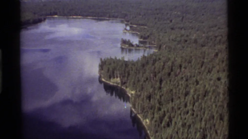 1972 8mm aerial footage over Holland Lake, showing dense Montana forest, calm water, and a narrow wooded point.