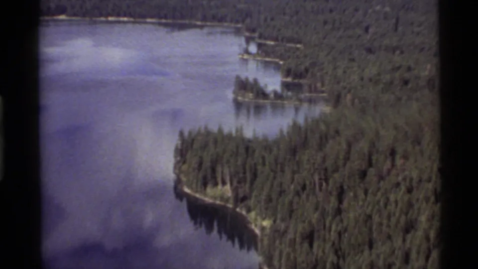 Aerial 8mm view of Holland Lake in 1972, with evergreen trees along a curved shore and open water filling the frame.