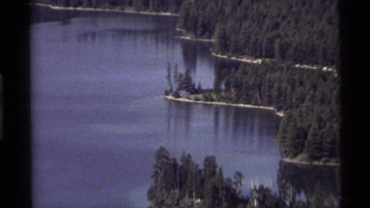 Holland Lake appears in a soft 8mm frame with dense forest, still water, and a tree-covered island.