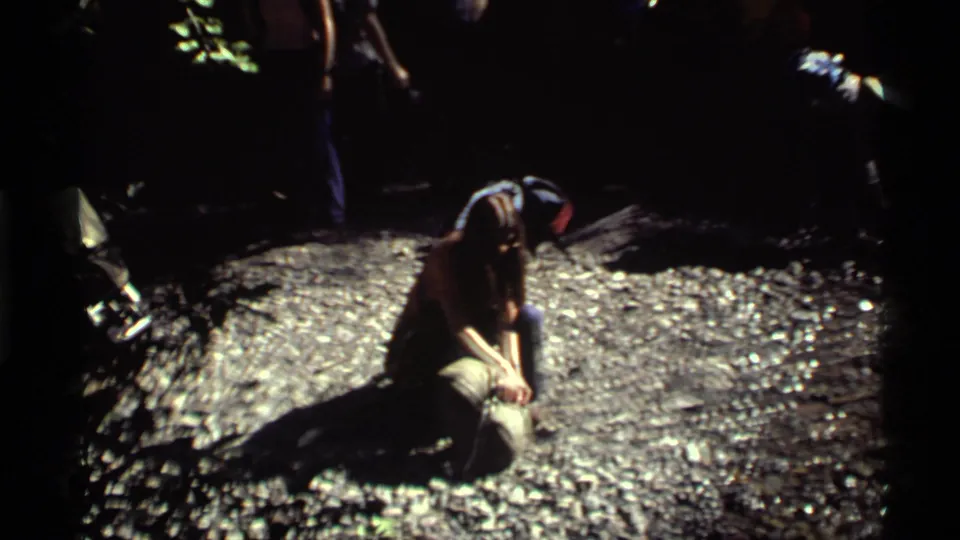 Dark 1972 home-movie campsite frame with a person kneeling on gravel while other figures stand in shadow.