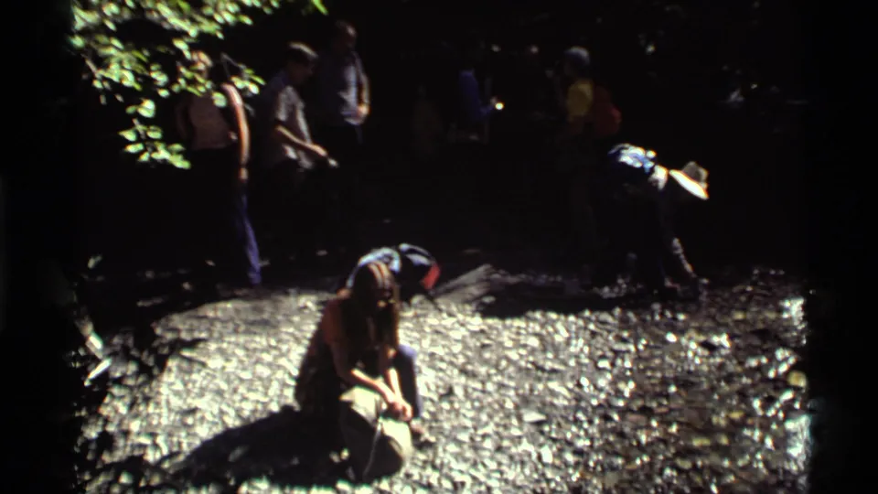 1972 8mm Holland Lake footage shows hikers in shade beside a rocky spring area as a woman kneels to drink.