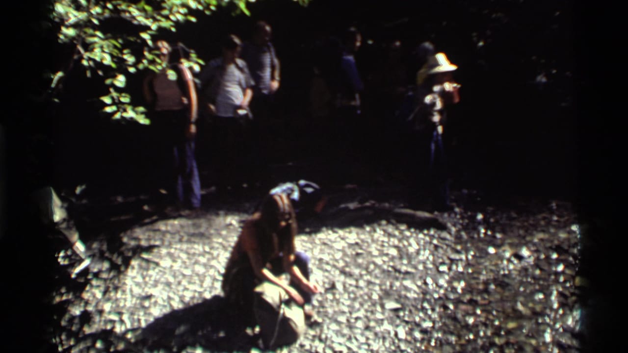 At Holland Lake, Montana, a kneeling hiker drinks near a rocky spring while the group waits under dappled forest light.