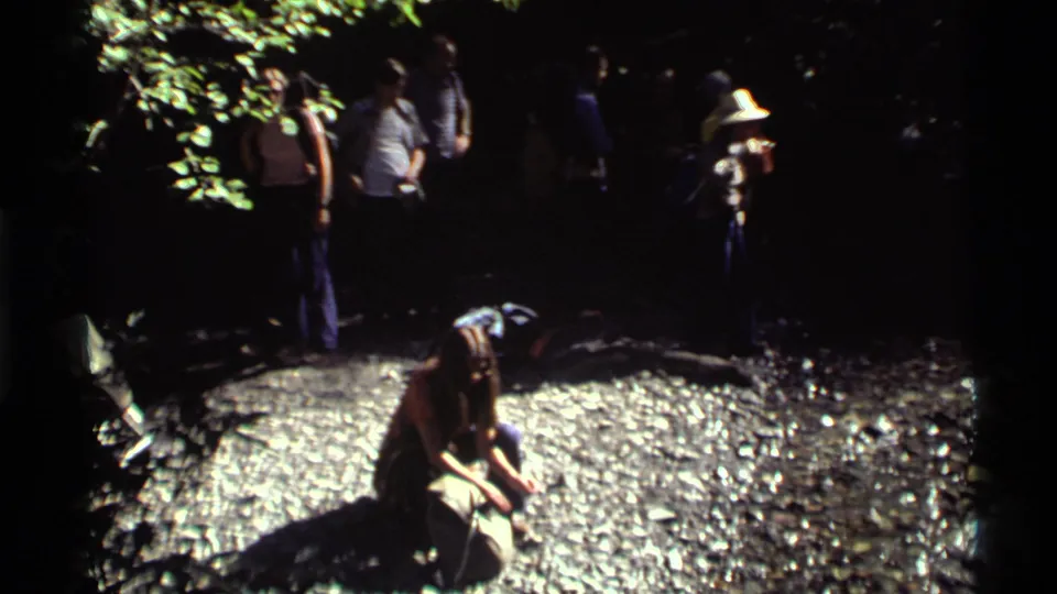 At Holland Lake, Montana, a kneeling hiker drinks near a rocky spring while the group waits under dappled forest light.
