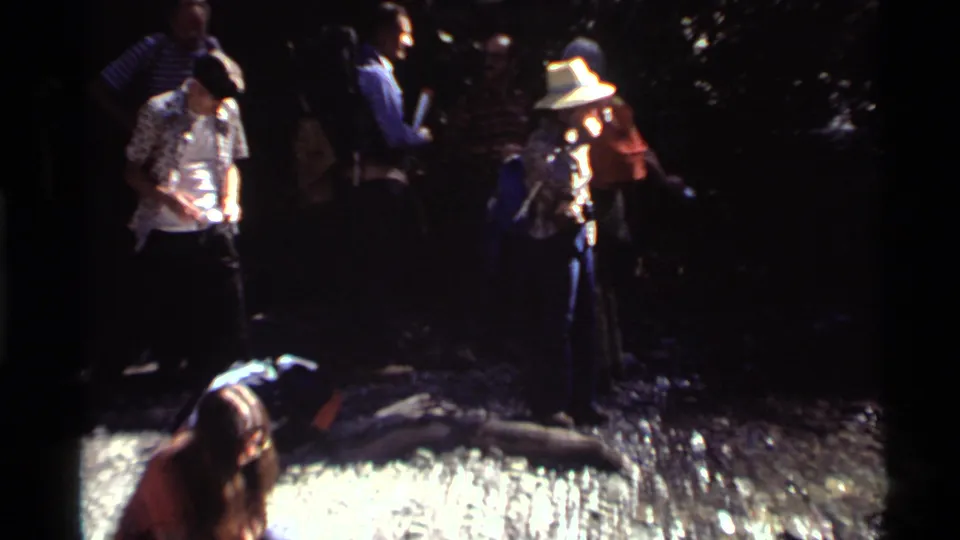The 1972 home-movie frame shows hikers gathered at the shaded streambank with backpacks, hats, and casual outdoor clothing.