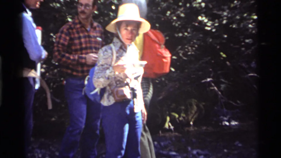 A woman in a wide-brim hat and blue trousers stands near the camera at Holland Lake, with other hikers and packs behind her.