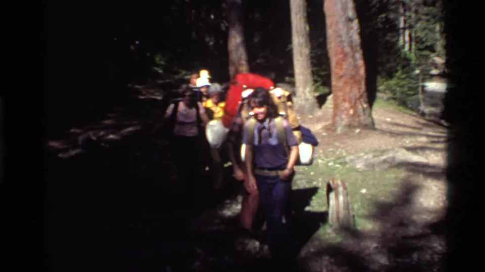 1972 Holland Lake, Montana 8mm frame showing hikers with packs moving through a sunlit and shadowed pine forest trail.