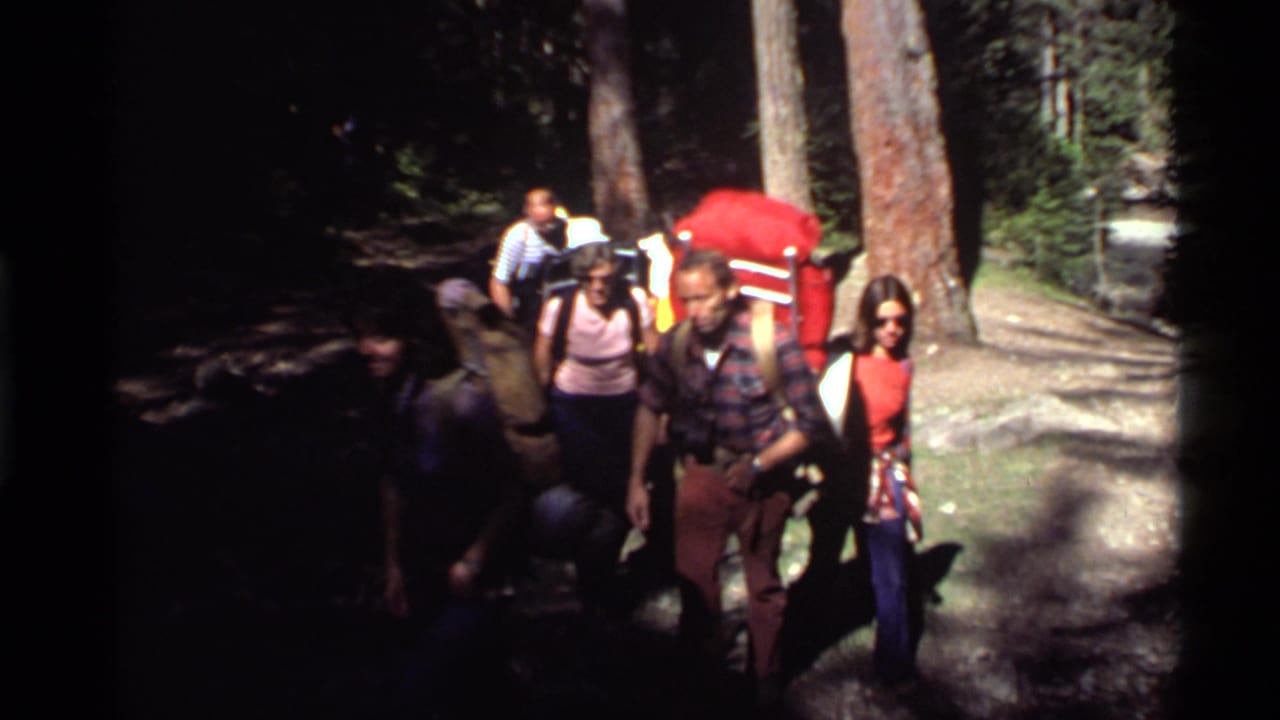 A mixed group of hikers in 1970s clothing walks past the camera on a shaded Holland Lake forest path.