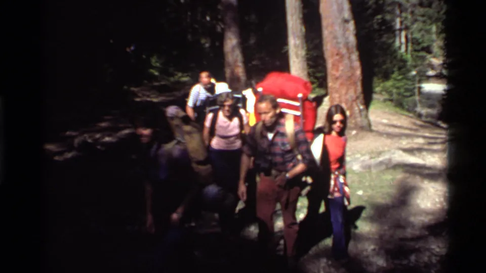 A mixed group of hikers in 1970s clothing walks past the camera on a shaded Holland Lake forest path.