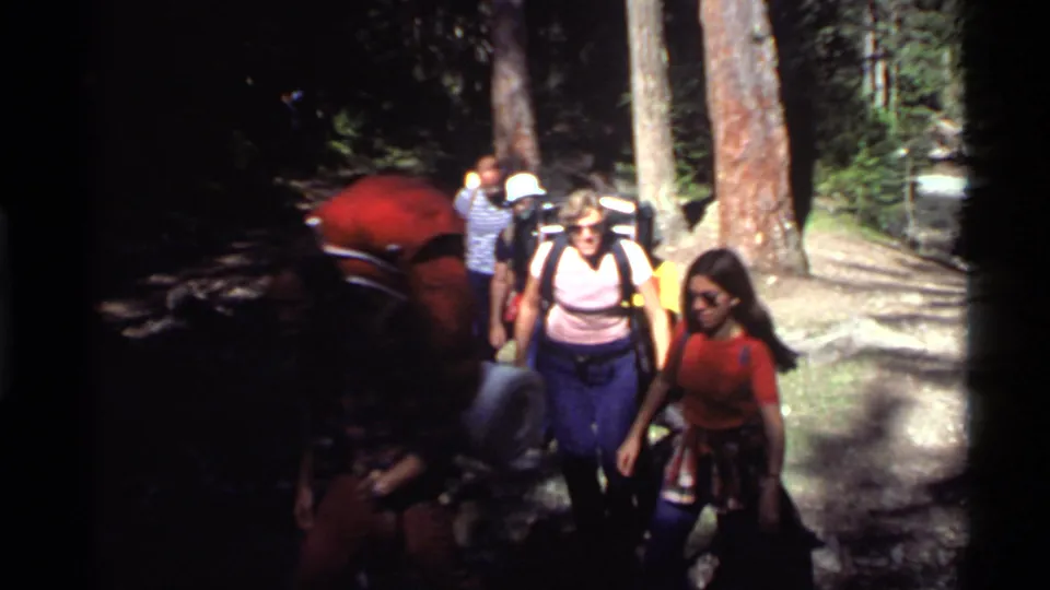 1970s hikers with backpacks and casual tops move close to the camera on a wooded trail in the Holland Lake area.