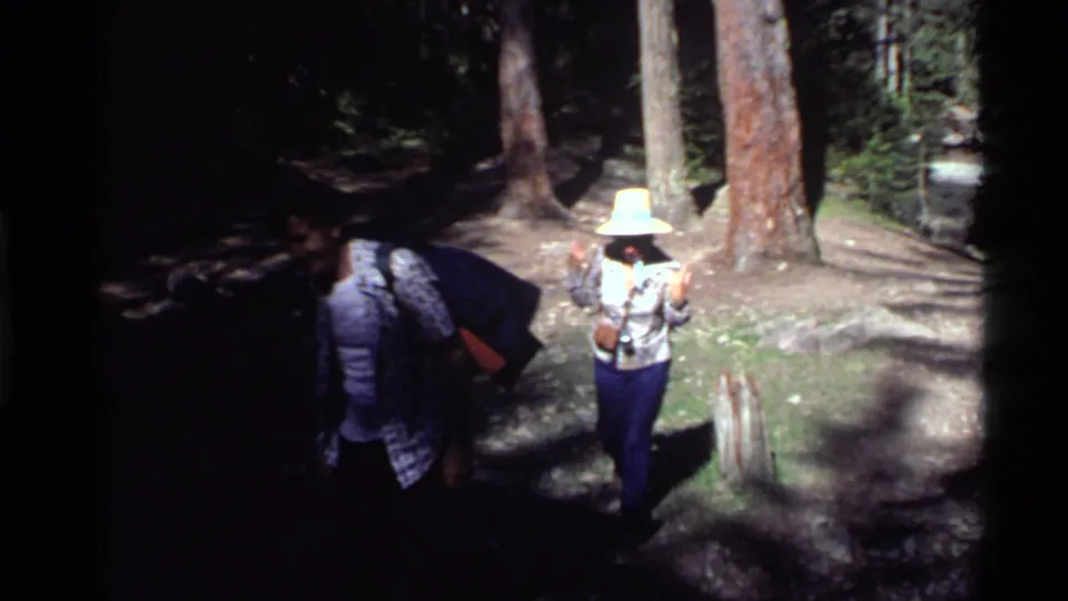 1972 Holland Lake home-movie frame showing hikers on a sun-dappled forest trail, including a woman in a wide-brim hat looking toward the camera.