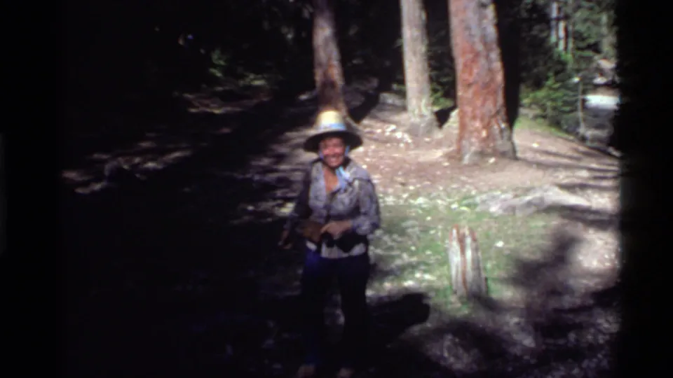 1972 Holland Lake frame of a smiling woman in a hat and patterned shirt on a shaded pine-forest trail.