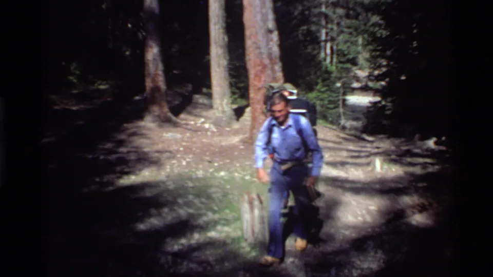 1972 Holland Lake home-movie frame showing an adult man with a backpack moving along a pine-forest trail.