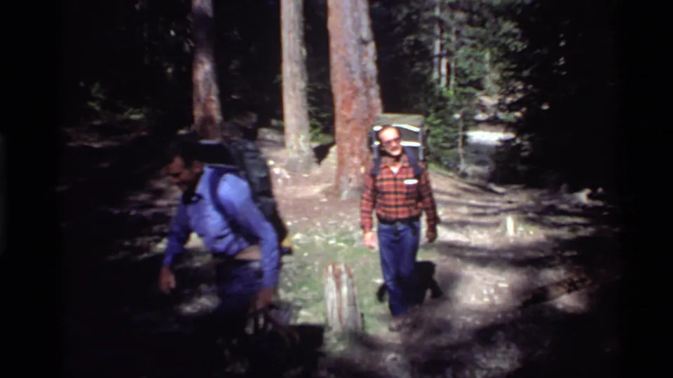 1972 Holland Lake frame showing two adult male hikers with backpacks on a shaded trail among tall pines.