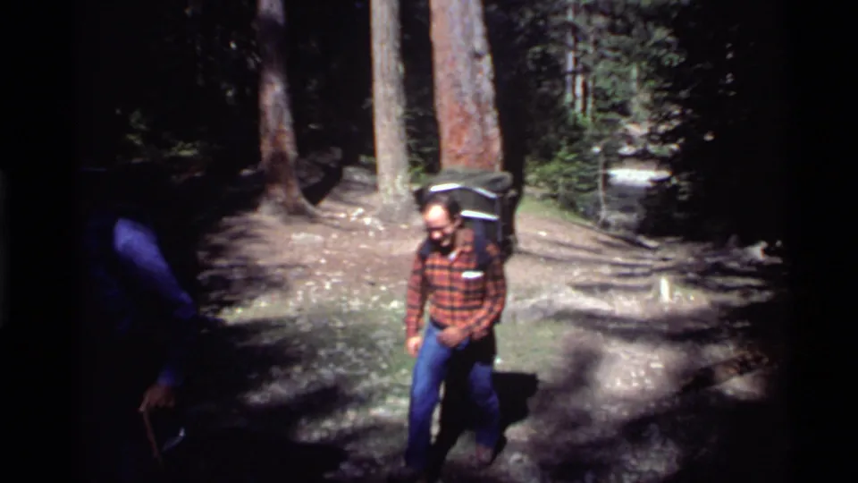 A hiker in a red checkered shirt and blue jeans walks with a backpack in the Holland Lake, Montana woods in 1972.