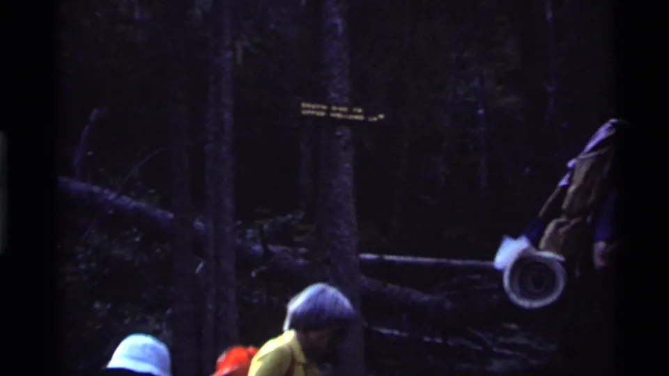 Backpacked hikers stand near a forest trail sign in the Holland Lake, Montana woods during the 1972 home movie sequence.