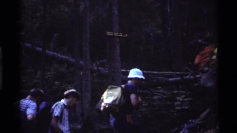Group of hikers in period clothing and packs moves along a wooded trail at Holland Lake, Montana, in 1972 footage.