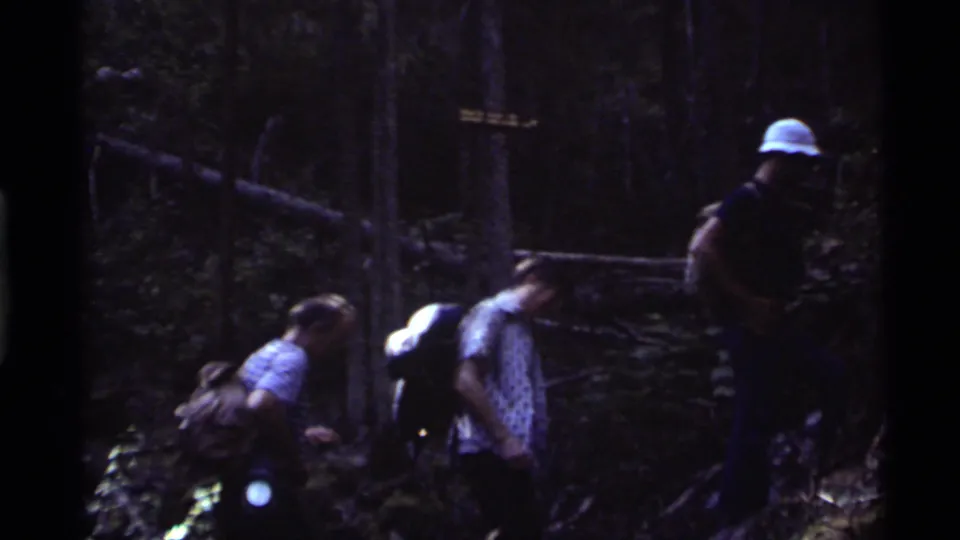 1972 8mm frame of hikers navigating uneven forest ground with packs and hats near Holland Lake, Montana.