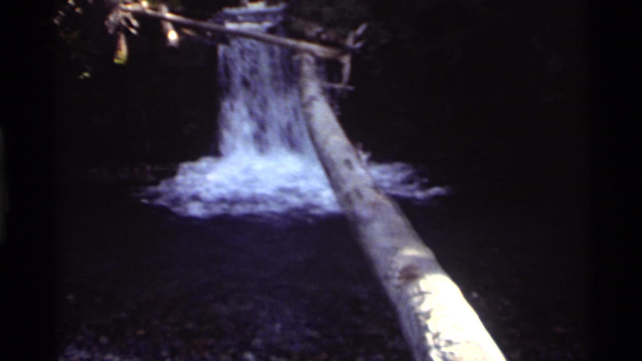 1972 8mm home-movie frame from Holland Lake, Montana, showing a shaded waterfall, white splash water, and a fallen log.