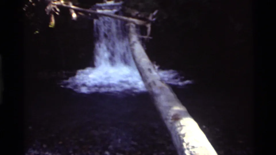 1972 8mm home-movie frame from Holland Lake, Montana, showing a shaded waterfall, white splash water, and a fallen log.