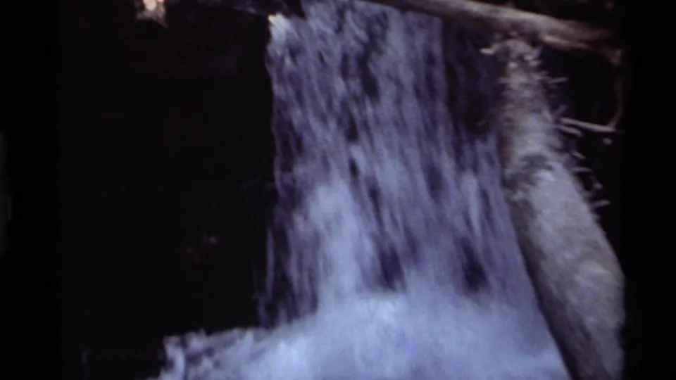 Close 8mm view of the Holland Lake waterfall, showing blurred falling water, a large log, and pale foam at the pool edge.