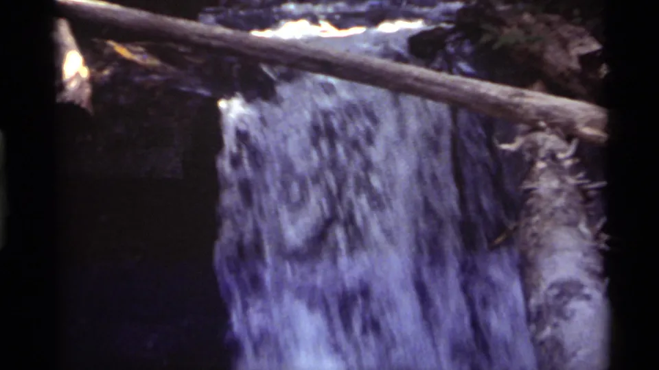 Shaded 1972 8mm frame of the Holland Lake waterfall with a horizontal fallen log crossing above the water.