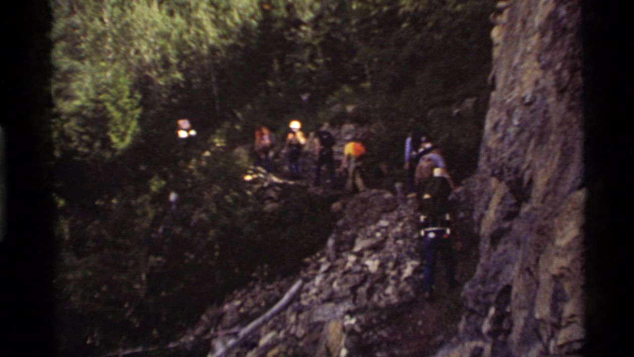 A group of hikers at Holland Lake, Montana, in 1972 moves along a narrow rocky forest trail with backpacks and hard hats visible.