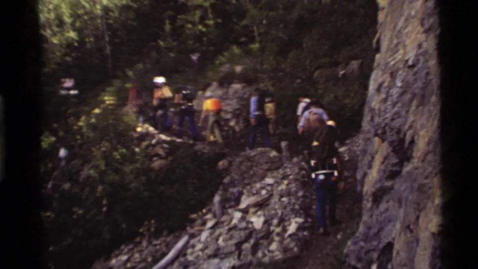 The group continues on a rocky Holland Lake forest path, with backpacks and light-colored hard hats visible among trees and boulders.