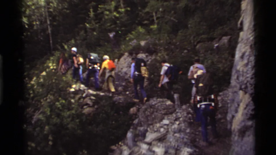 Backpackers in 1972 Holland Lake footage walk away from the handheld camera along a dirt and rock trail through dense forest.