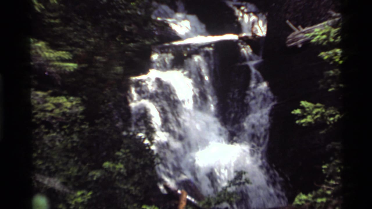 A waterfall drops through forested rocks in the Holland Lake, Montana sequence, framed in handheld 8mm footage.