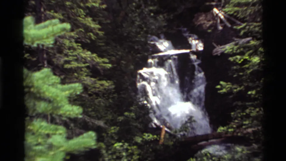 The 1972 Montana home-movie clip shows the waterfall from farther back, with foreground evergreen branches and rocky terrain.