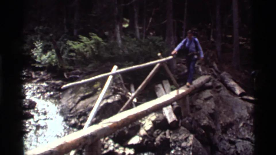 At Holland Lake in 1972, an adult hiker steps across a simple log bridge over rushing water and rocks.