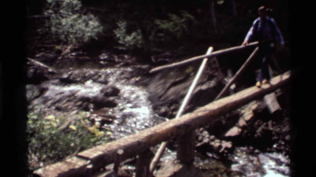 1972 home-movie frame from Holland Lake, Montana, showing an adult hiker balancing on a log bridge above a rocky stream.