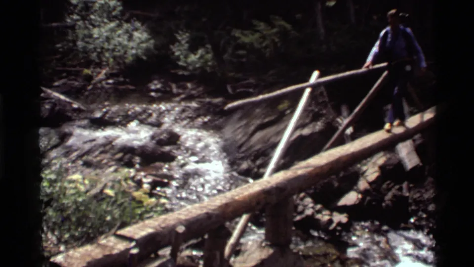 1972 home-movie frame from Holland Lake, Montana, showing an adult hiker balancing on a log bridge above a rocky stream.
