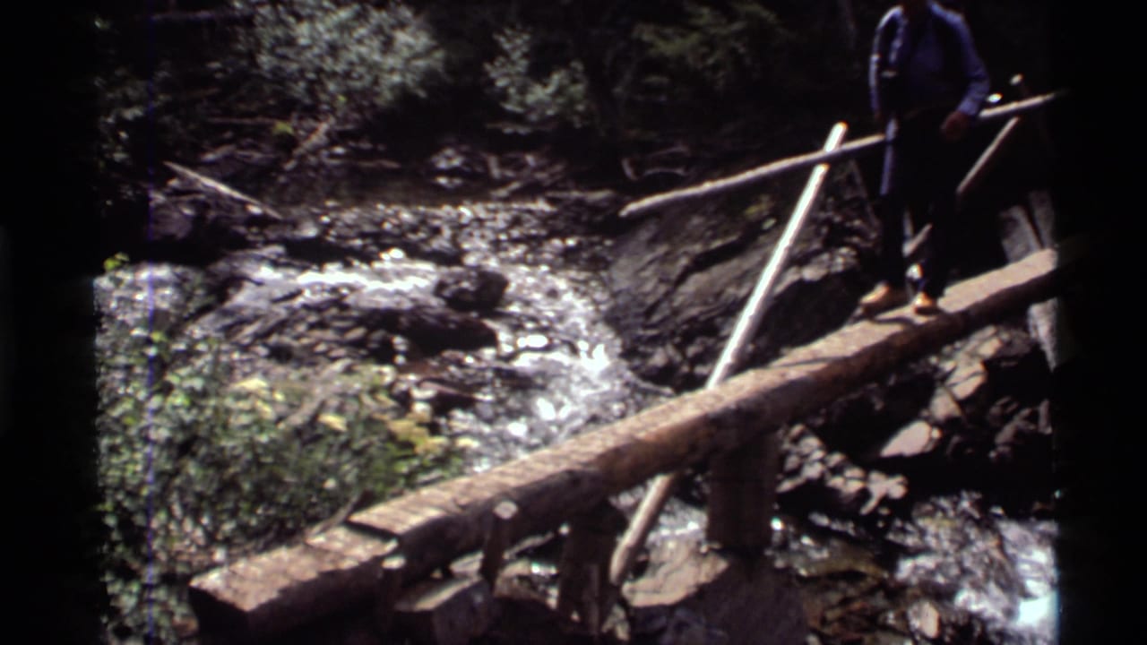 1972 Holland Lake hiking footage shows a man on a rustic log footbridge with trees, rocks, and moving water below.