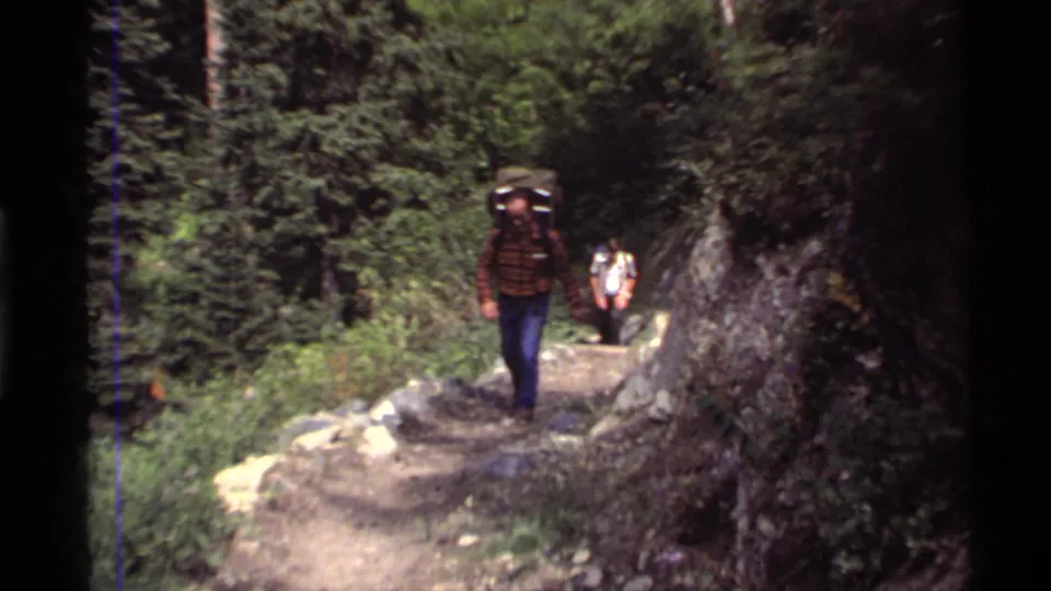 1972 home-movie view of two adult men hiking a narrow trail at Holland Lake, Montana, with backpacks and dense foliage around them.