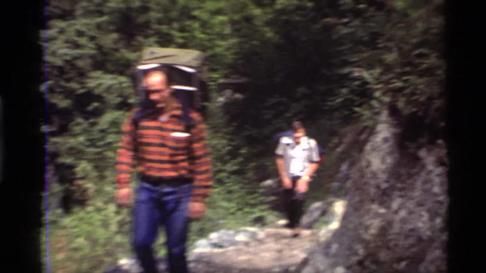 1972 Holland Lake frame with two adult hikers approaching the camera on a forest trail, one blurred in the foreground with a backpack.