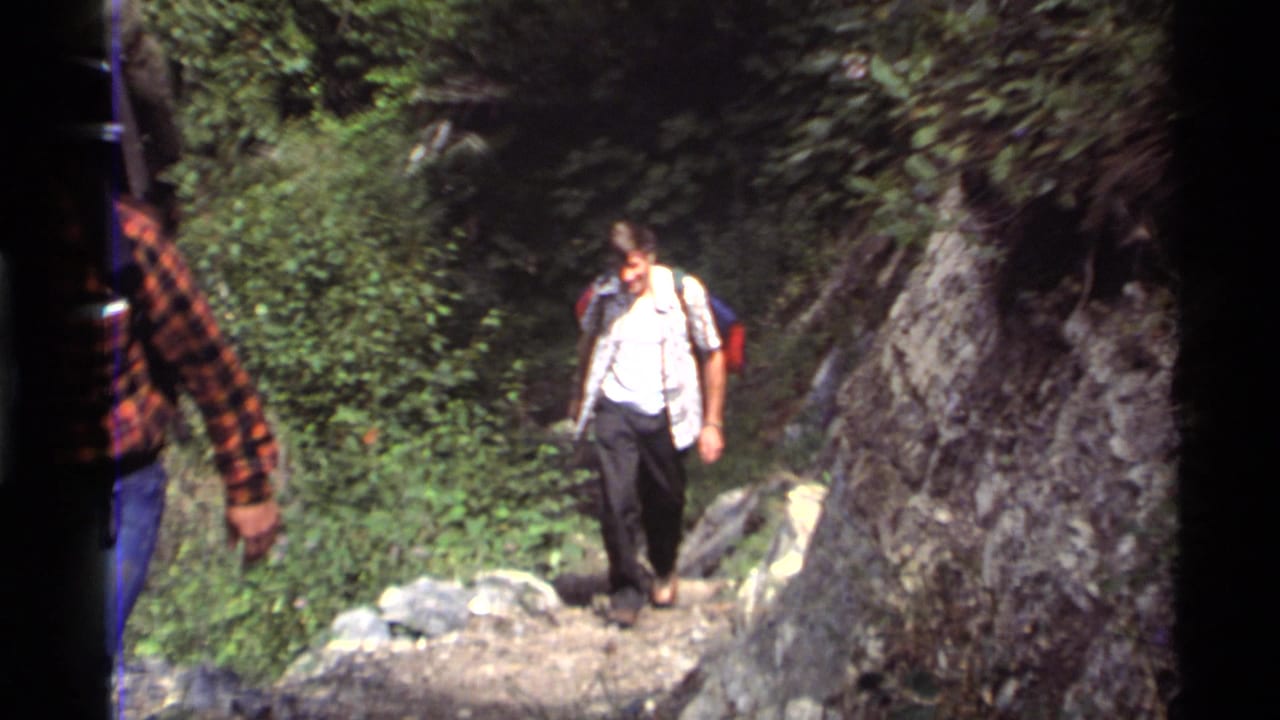 1972 Holland Lake, Montana 8mm frame showing men with backpacks walking down a rocky forest trail through dense brush.