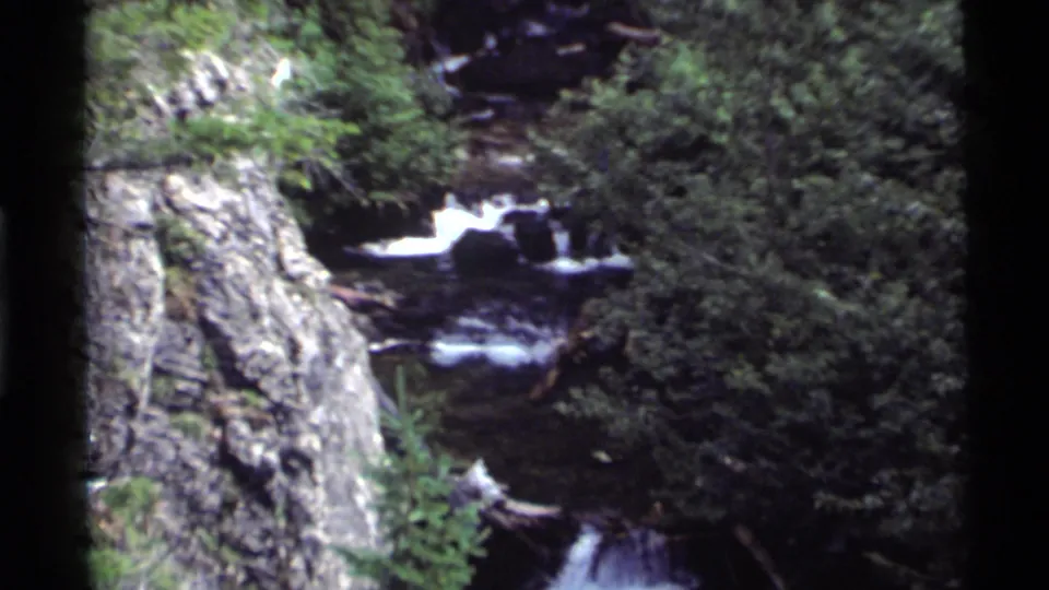 A fast rocky stream flows through dense green forest near Holland Lake, Montana, captured on 1972 8mm home-movie film.
