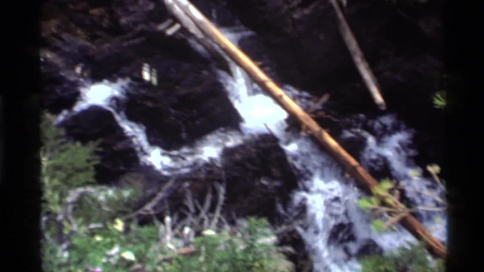 1972 Holland Lake 8mm frame showing water cascading over rocks, crossed by a narrow fallen trunk.
