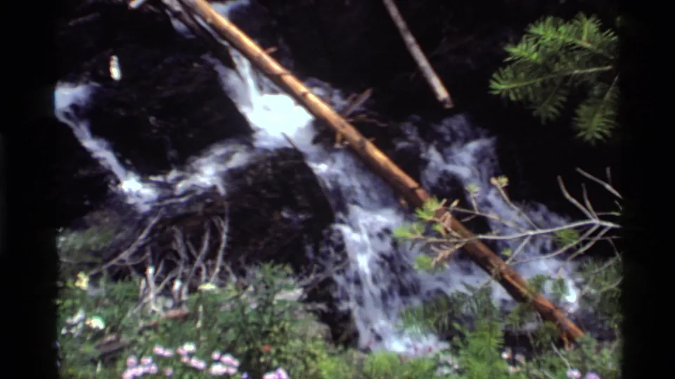 1972 Montana home-movie frame of a rocky forest cascade with fallen wood and evergreen branches.