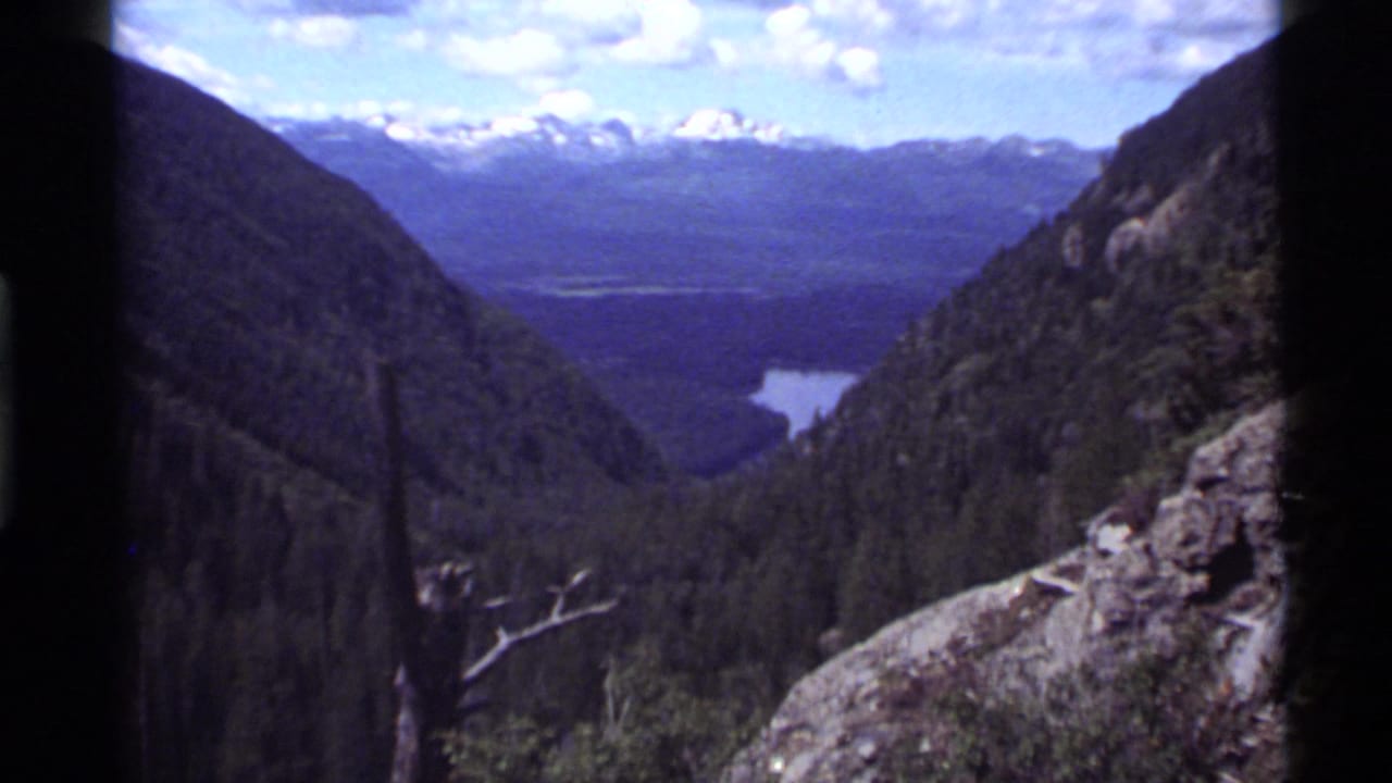 1972 8mm view near Holland Lake, Montana, showing forested slopes, a distant lake, rocky foreground, and snowy peaks.