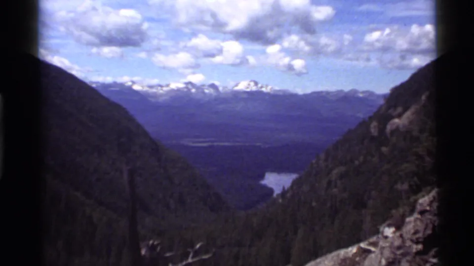 1972 8mm frame from Holland Lake, Montana, showing a broad forested valley, distant water, and snow-bright peaks under clouds.
