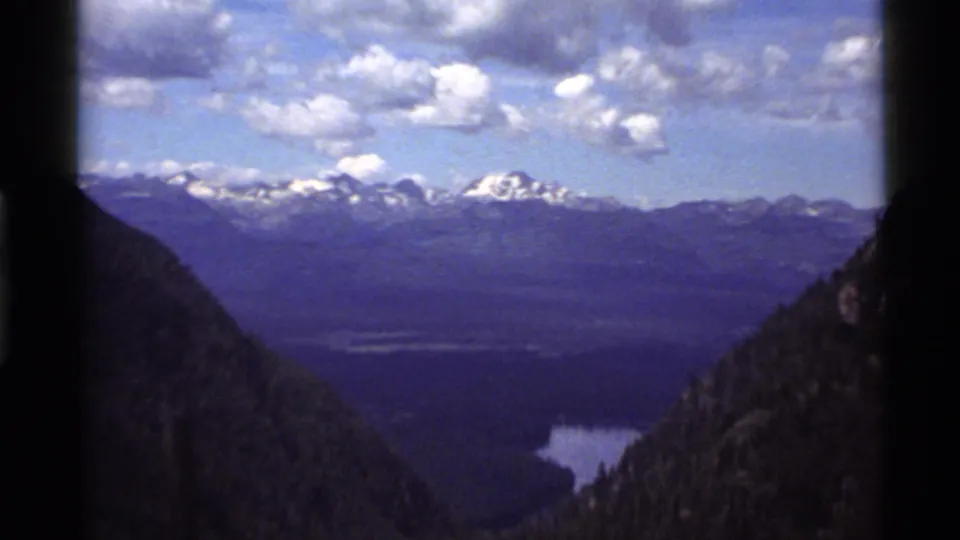 1972 8mm Holland Lake, Montana view with snowy mountain peaks, forested valley floor, lake water, and dark frame-edge shadows.