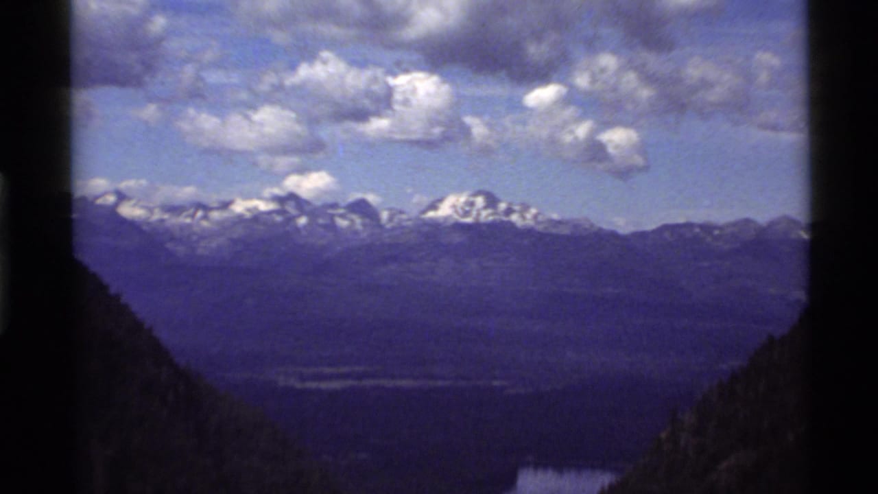 1972 Holland Lake, Montana home-movie frame showing distant snow-covered mountains, dark slopes, lake water, and scattered clouds.
