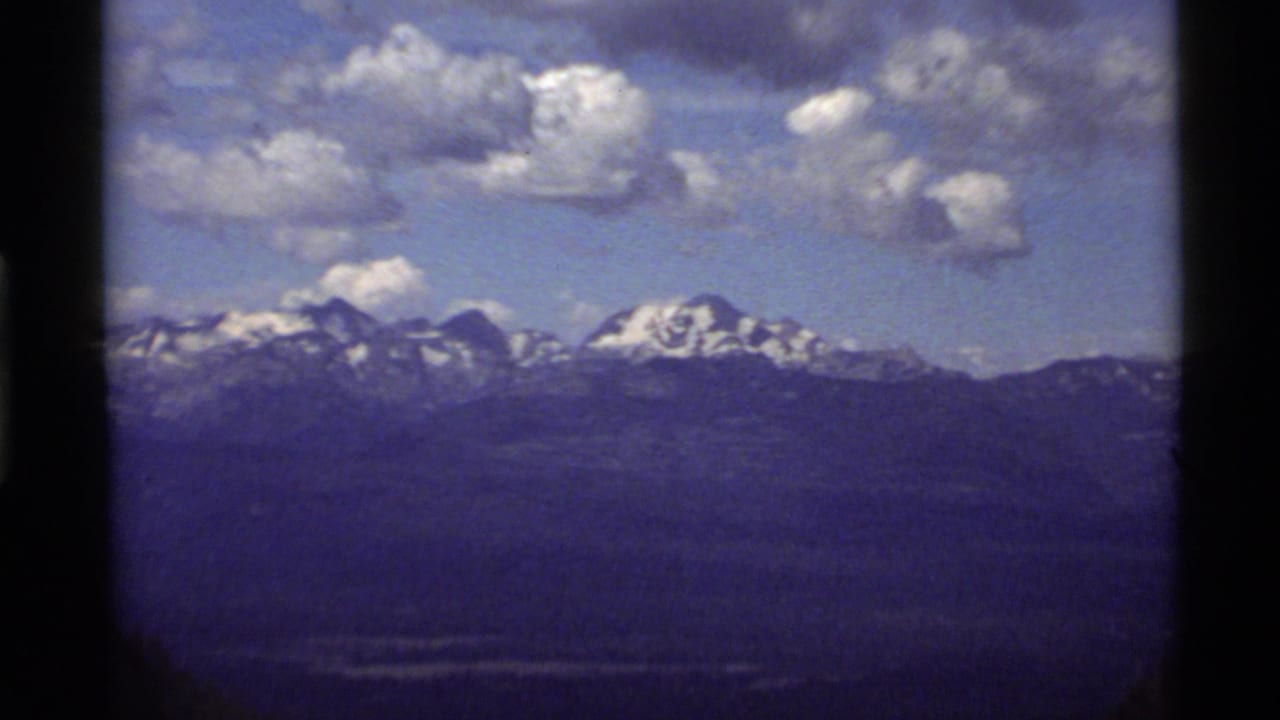Snowy mountain range under scattered clouds with dark forested land in the foreground.