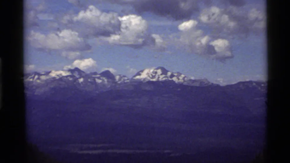 1972 Holland Lake, Montana frame with snow-covered peaks, blue-purple distant range, forested foreground, and cloudy sky.