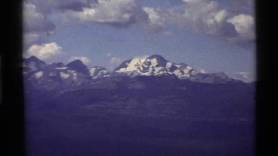Closer 1972 Holland Lake, Montana home-movie view of snowy peaks and layered dark ridges under scattered clouds.