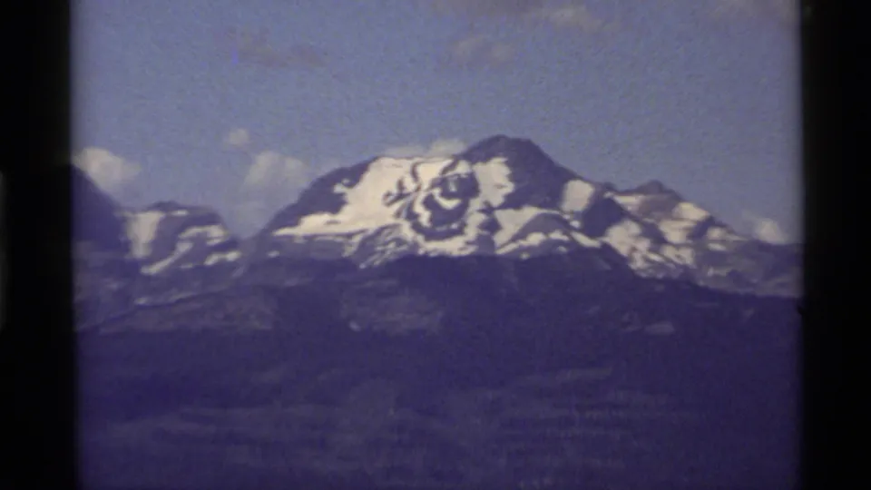 Zoomed 1972 Holland Lake, Montana frame isolating a snow-covered peak, dark lower ridges, and pale clouds.