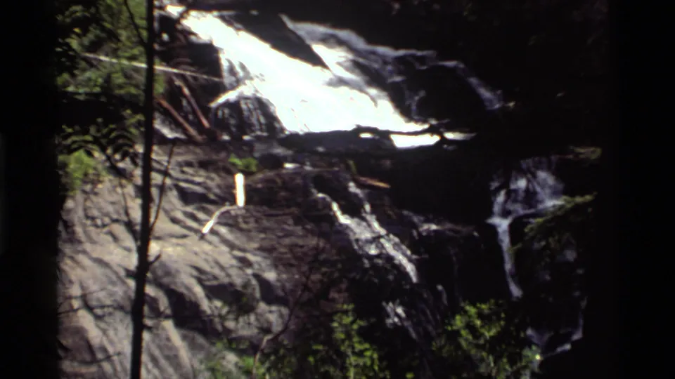 1972 Holland Lake, Montana waterfall view with bright water, dark rock ledges, and surrounding trees.