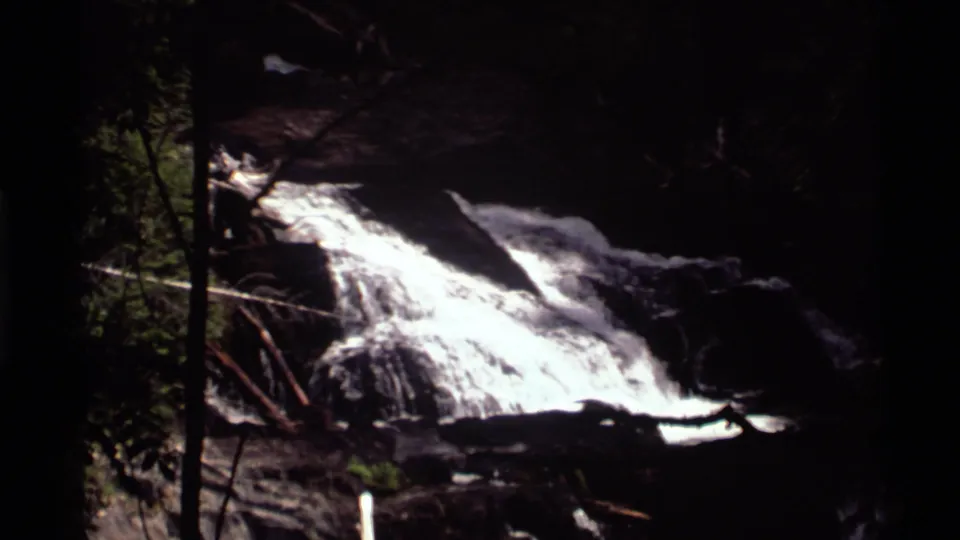 1972 Holland Lake, Montana 8mm frame showing foamy water descending over a rock slope bordered by forest growth.
